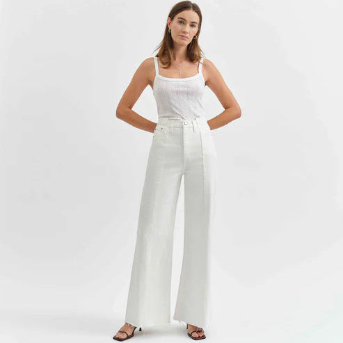 Photograph of a model in a studio wearing white high-waisted wide-leg jeans with a distinctive vertical front seam, a white ribbed tank top, and strappy black sandals