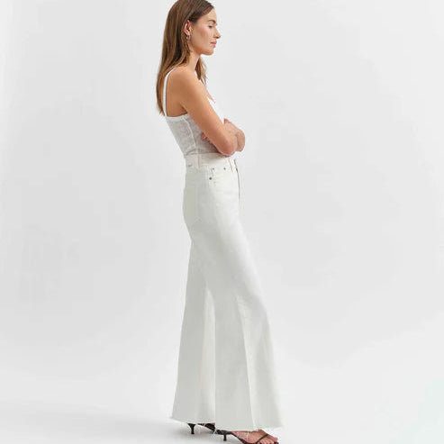 Studio photograph of a woman modeling white high-waisted flared jeans and a ribbed tank top in a minimalist studio featuring raw frayed hems and black strappy kitten heels