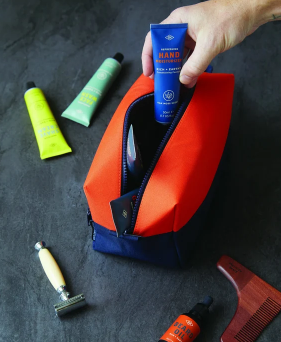 Hand placing a tube of hand cream into an open orange and navy toiletry bag beside a razor and comb