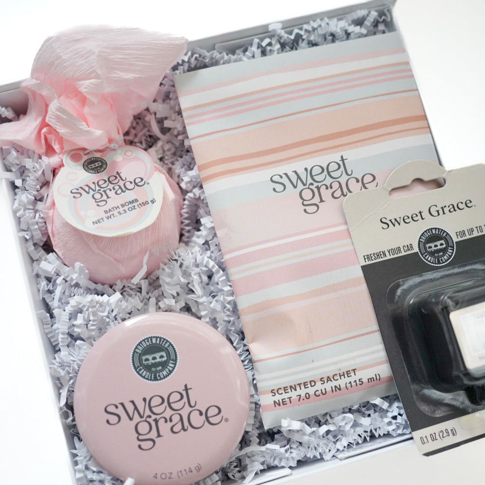 Commercial product photography of Bridgewater Candle Company featuring a Sweet Grace fragrance gift set in a box with a pink-wrapped bath bomb, a striped scented sachet, and a round pink tin resting on white crinkle-cut paper