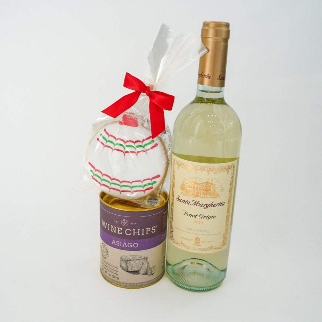 Studio photograph of a gourmet gift set featuring a bottle of Santa Margherita Pinot Grigio 2024, a purple tin of Asiago Wine Chips, and a large iced cookie decorated with red and green scalloped patterns and a bright red ribbon bow against a white background