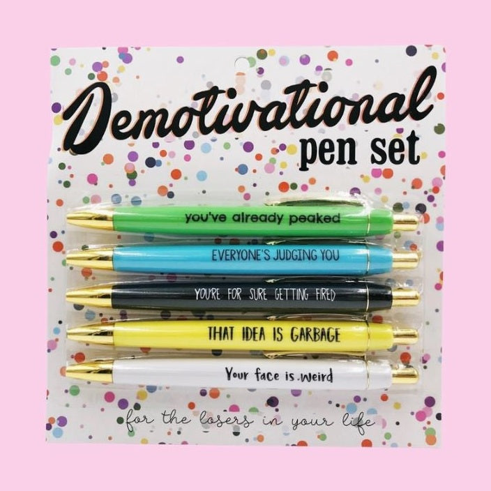 Product photograph of a Demotivational pen set on a pink background featuring five gold-accented pens in green, blue, black, yellow, and white with cynical phrases against a vibrant confetti-patterned package labeled for the losers in your life