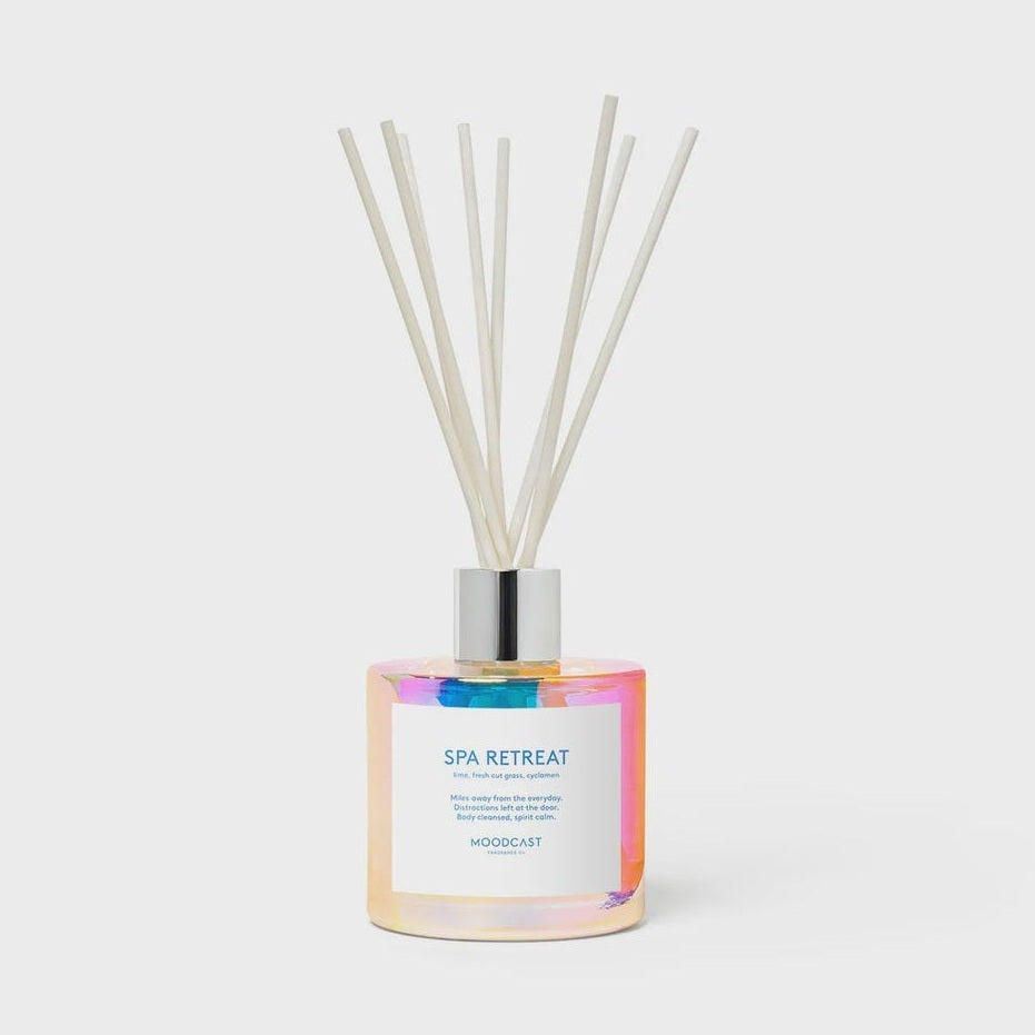 Product photograph by Moodcast of a Spa Retreat reed diffuser featuring an iridescent holographic glass bottle with a silver metallic cap and fanned-out white reeds against a neutral studio background