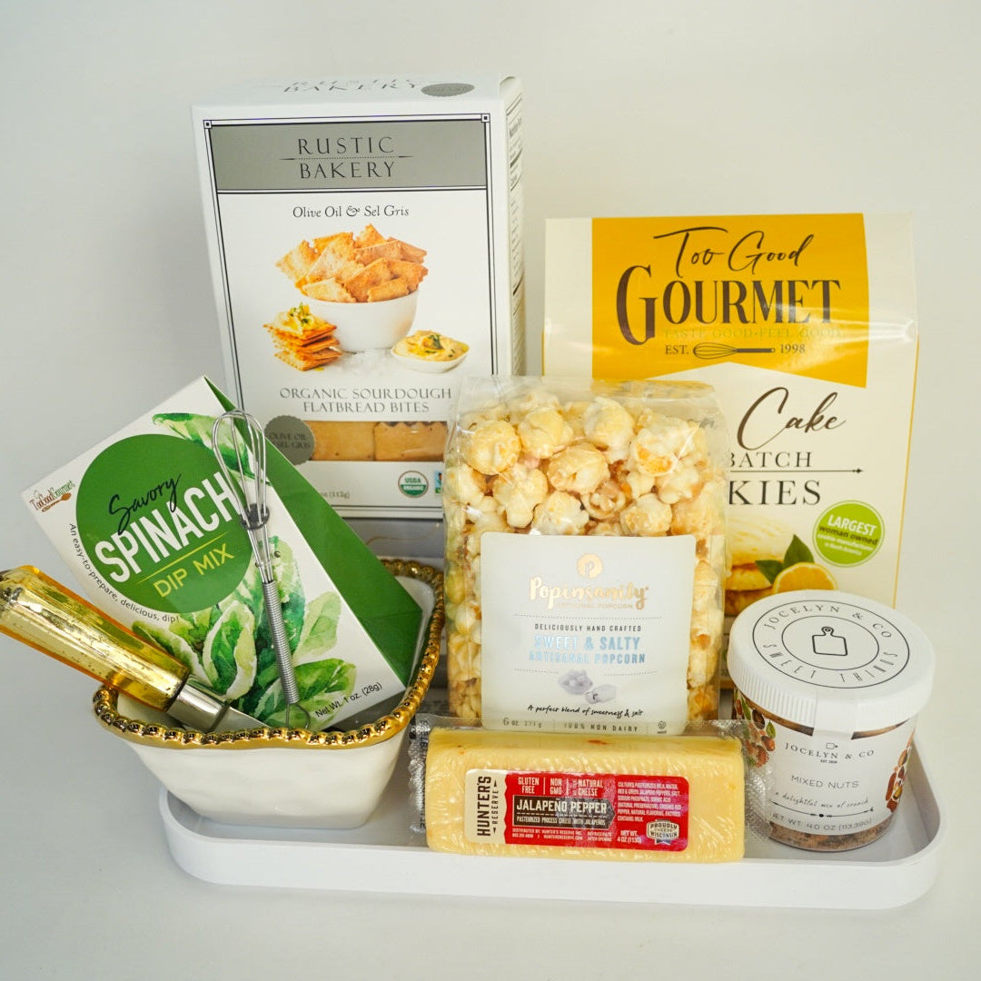 Product photograph of a gourmet food gift set on a white tray featuring packaging for Rustic Bakery and Popinsanity alongside a vibrant green spinach dip box, a block of yellow jalapeno cheese, and a small gold-rimmed bowl containing a miniature metal whisk