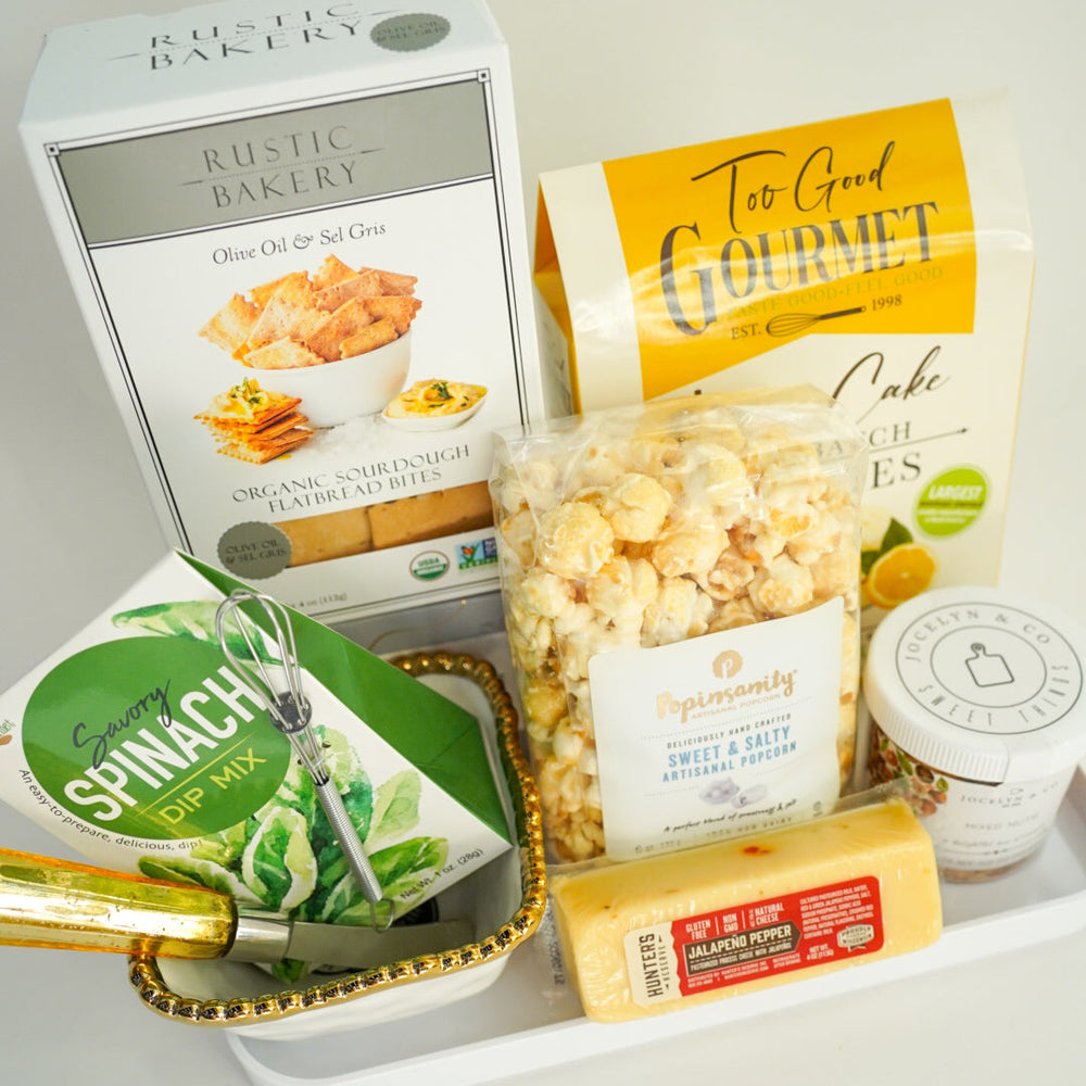 Photograph of a curated gourmet food gift set featuring Rustic Bakery flatbread bites and Popinsanity popcorn arranged in a white tray with a gold-rimmed decorative bowl and a block of yellow jalapeño pepper cheese