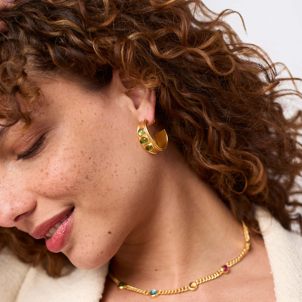 Close-up photograph of a woman with freckled skin and curly auburn hair showcasing a wide gold hoop earring with three green cabochons and a gold chain necklace with vibrant multi-colored gems