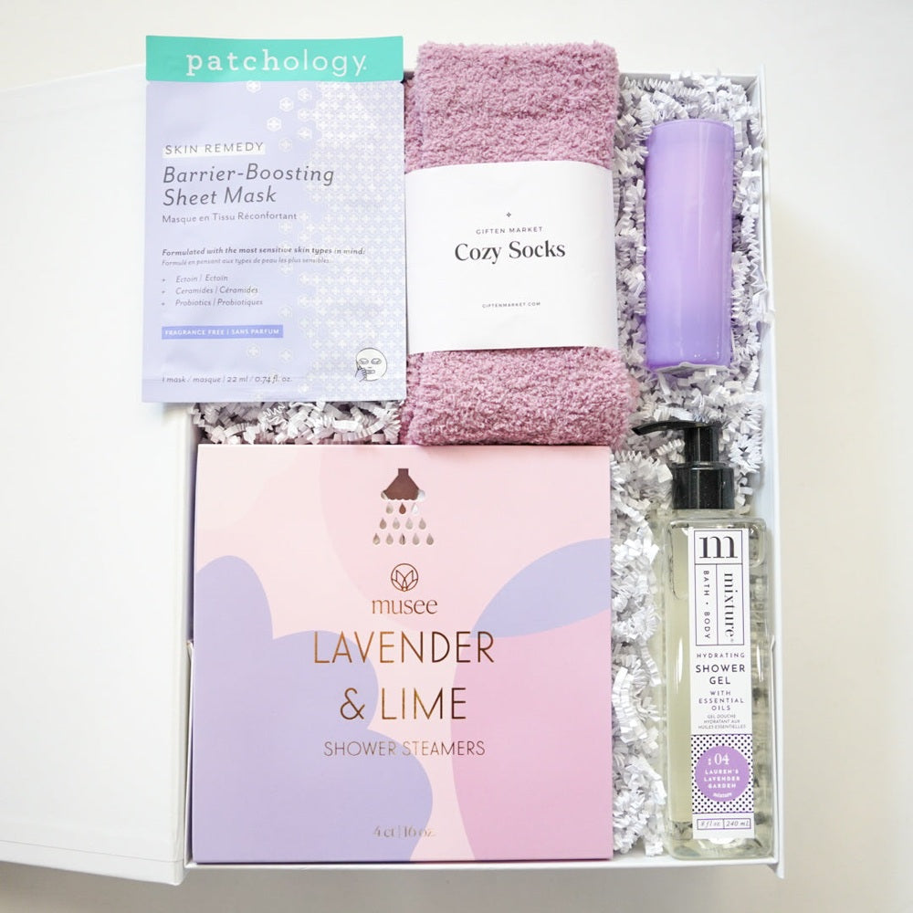Photograph by Giften Market of a curated self-care gift box containing a Patchology sheet mask, plush mauve cozy socks, and Musee lavender shower steamers nestled in white crinkle paper alongside a tall purple tumbler and clear shower gel bottle