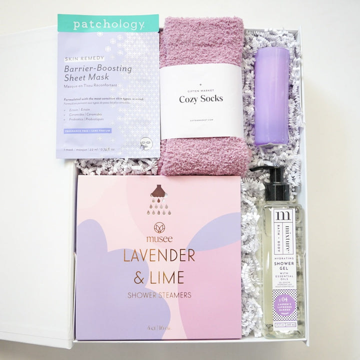 Photograph by Giften Market of a curated self-care gift box containing a Patchology sheet mask, plush mauve cozy socks, and Musee lavender shower steamers nestled in white crinkle paper alongside a tall purple tumbler and clear shower gel bottle