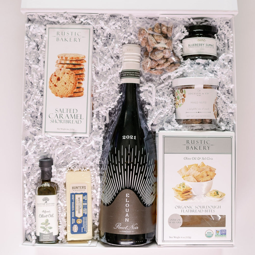 Studio photograph of a curated gourmet gift box featuring a bottle of 2021 Elouan Pinot Noir, boxes of Rustic Bakery artisanal crackers, and a block of Hunter&