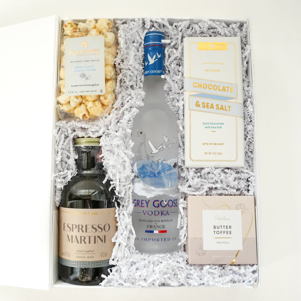Commercial photograph of a curated gourmet gift box featuring a frosted bottle of Grey Goose vodka with its iconic flying geese logo, a dark Espresso Martini cocktail mixer, and artisanal sweet and salty popcorn nestled in white shredded paper filler inside a clean white container