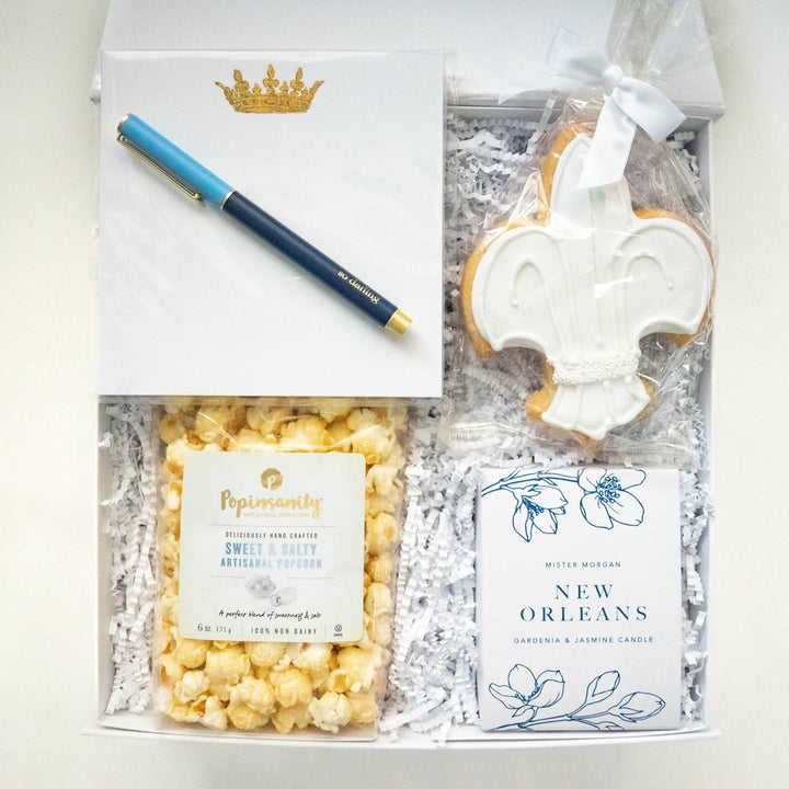 Flat lay photograph of a New Orleans themed gift box featuring a large white-iced fleur-de-lis cookie, a gold crown-embossed notepad with a navy blue pen, and a bag of Popinsanity sweet and salty popcorn nestled in white shredded paper