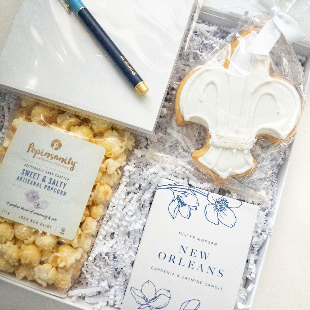 Photograph of a New Orleans-themed curated gift box featuring an iconic white-iced fleur-de-lis cookie, Popinsanity sweet and salty popcorn, and a Mister Morgan gardenia candle paired with a navy blue pen with gold lettering on white crinkle paper