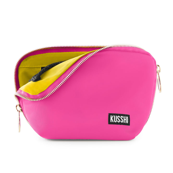 Product photography of a Kusshi makeup bag featuring a vibrant hot pink exterior, a contrasting bright yellow interior lining, and gold-toned zippers with a black and white brand label against a white background