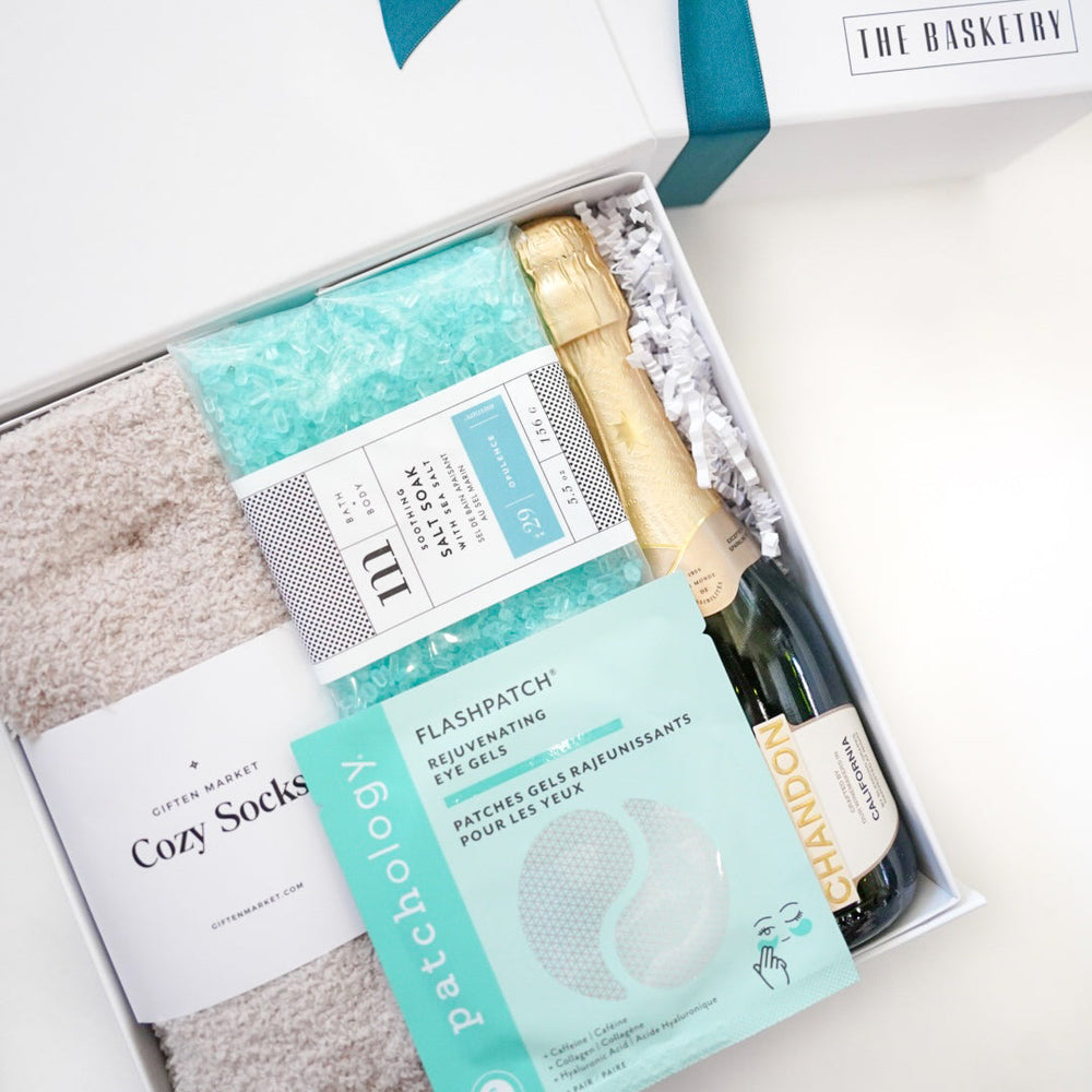 Gift set with Cozy Socks, Flax Patch, and a bottle of champagne in a white box with a teal ribbon.