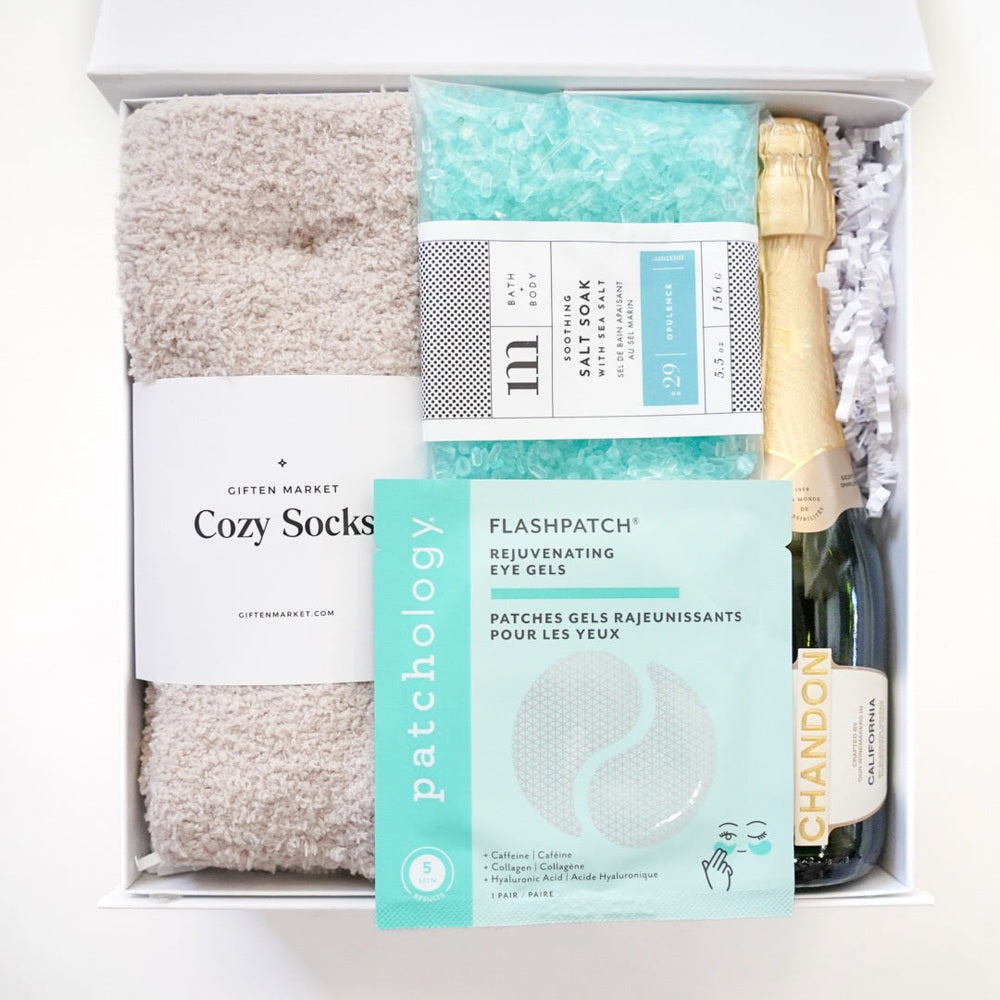 Studio photograph by Giften Market of a curated spa gift box featuring tan fuzzy socks, vibrant teal sea salt soak, Patchology eye gels, and a mini bottle of Chandon champagne arranged in a white box