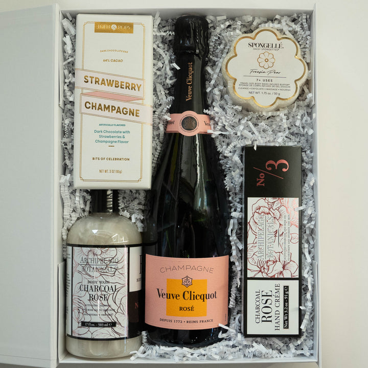 Photograph of a luxury gift box featuring a central bottle of Veuve Clicquot Rosé Champagne with a salmon-pink label nestled in white shredded paper alongside Archipelago Botanicals skincare and a flower-shaped Spongellé buffer