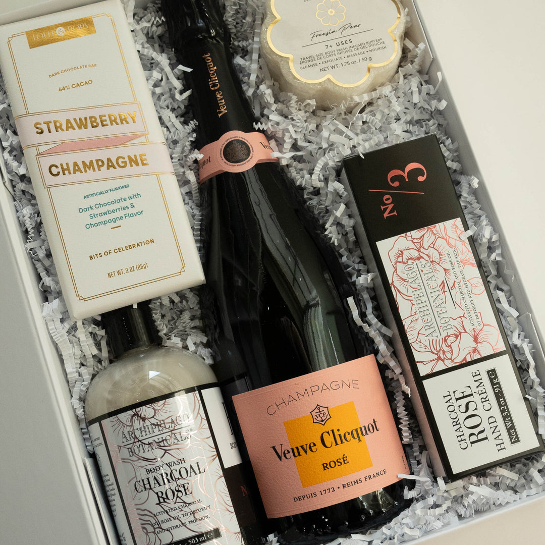 Photograph of a luxury gift box featuring a bottle of Veuve Clicquot Rosé Champagne, Archipelago Botanicals Charcoal Rose bath products, and a strawberry champagne chocolate bar nestled in white crinkle paper with peach-colored labels and gold accents