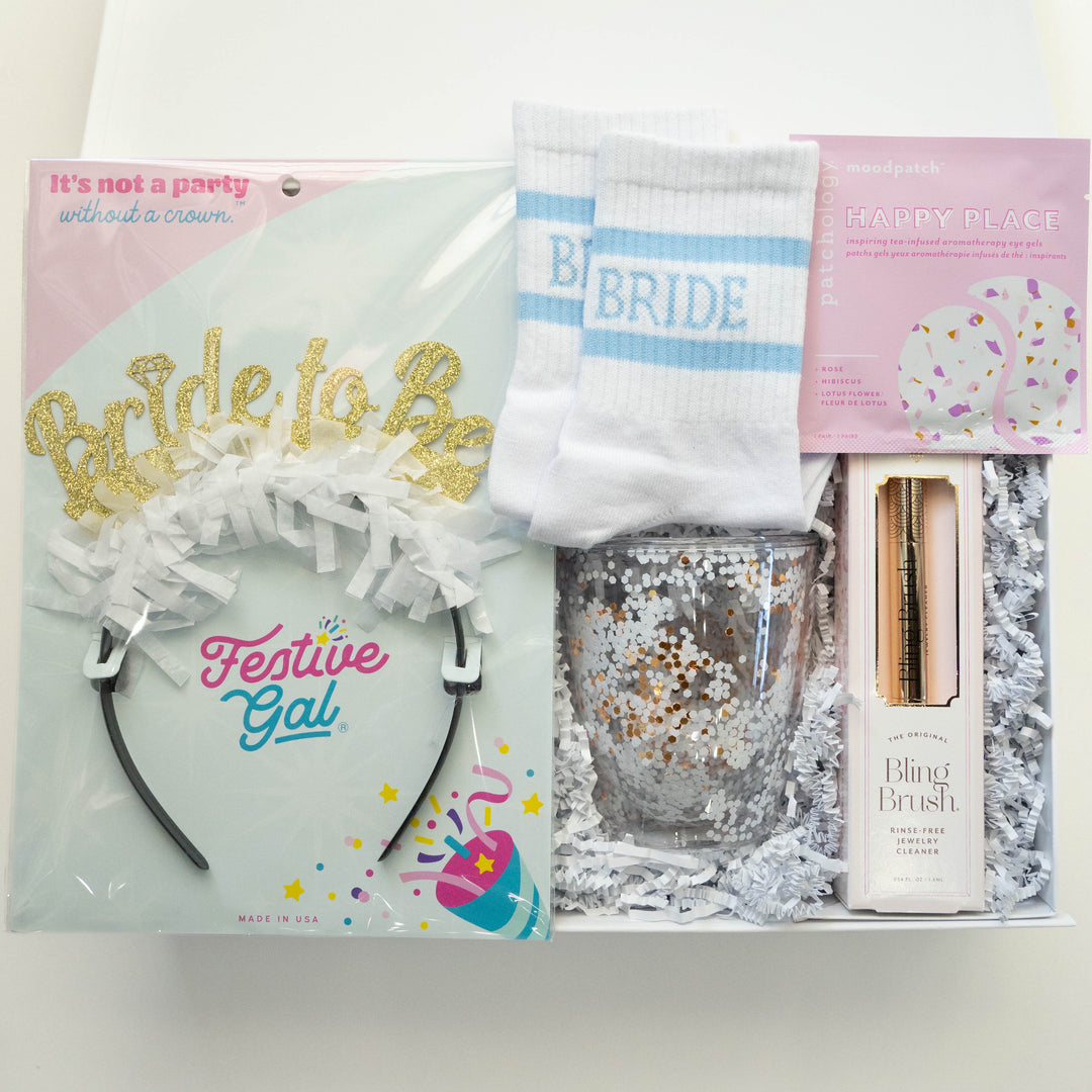 Photograph of a bridal gift box by Festive Gal featuring a gold glitter Bride to Be headband, white socks with light blue stripes, and a sparkling gold confetti tumbler nestled in white shredded paper