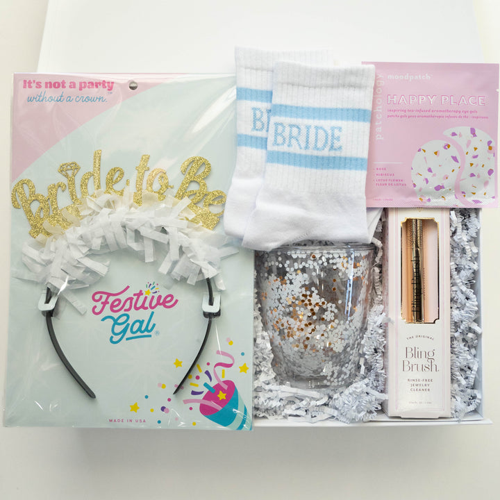 Photograph of a bridal gift box by Festive Gal featuring a gold glitter Bride to Be headband, white socks with light blue stripes, and a sparkling gold confetti tumbler nestled in white shredded paper