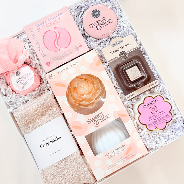 Professional product photograph of a Giften Market spa gift box featuring Sweet Grace and Spongellé brand products, a peach sola wood flower diffuser, pink Pathology eye gels, and fuzzy tan socks nestled in white crinkle paper