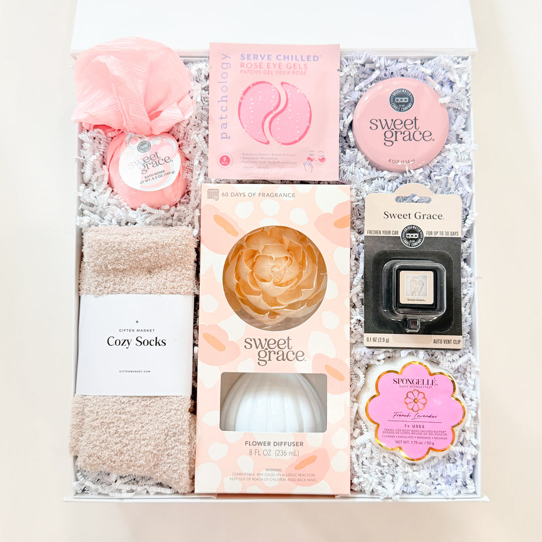 Photograph of a Giften Market self-care gift set inside a white box featuring a Sweet Grace flower diffuser, Patchology eye gels, fuzzy beige socks, and a peach-colored wooden flower on white shredded paper