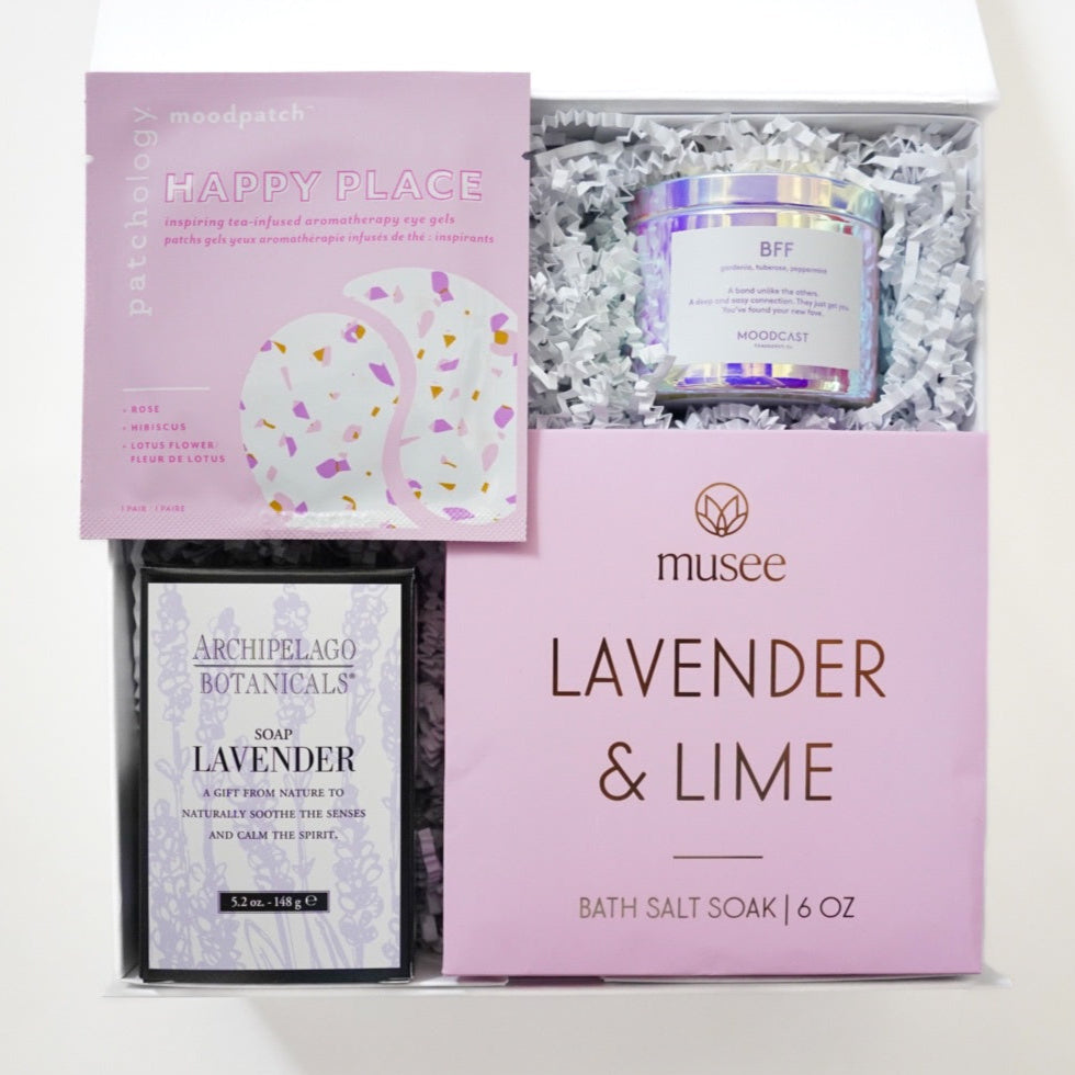 Photograph of a curated spa gift box featuring a pink Patchology eye gel packet, an iridescent Moodcast candle, and a Musee Lavender & Lime bath salt soak box nestled in white shredded paper filler