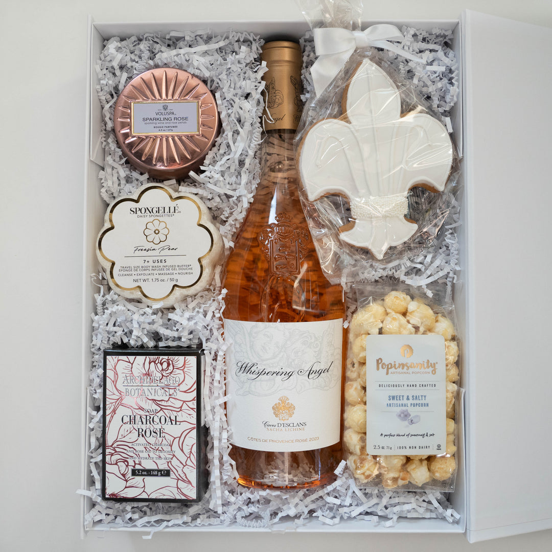 Photograph of a luxury curated gift box featuring a central bottle of Whispering Angel Rosé wine, a large white icing fleur-de-lis cookie, a rose gold Voluspa candle tin, and a flower-shaped Spongellé buffer nestled in white shredded paper filler
