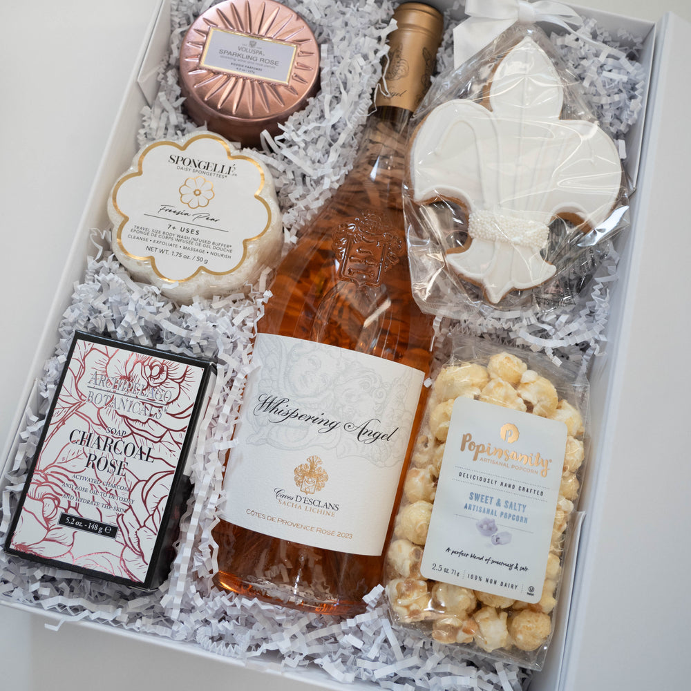 Photograph of a curated luxury gift box featuring a bottle of Whispering Angel Rosé, a rose-gold Voluspa candle tin, and a white fleur-de-lis frosted cookie nestled in white shredded paper filler