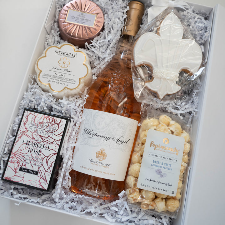 Photograph of a curated luxury gift box featuring a bottle of Whispering Angel Rosé, a rose-gold Voluspa candle tin, and a white fleur-de-lis frosted cookie nestled in white shredded paper filler
