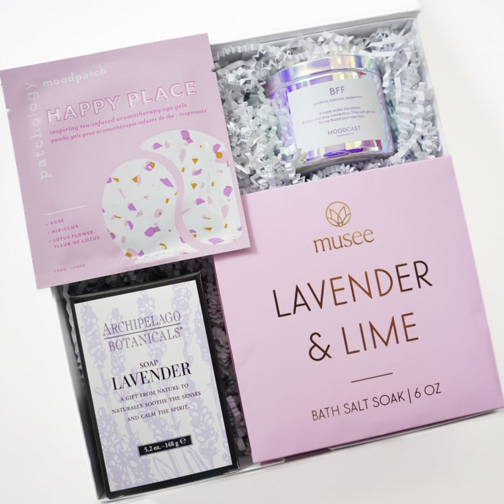 Digital photograph of a curated wellness gift box featuring Musee Lavender and Lime bath salts, Archipelago Botanicals lavender soap, and Patchology eye gels nestled in white crinkle paper beside a shimmering iridescent candle jar