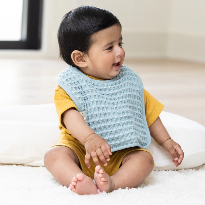 Photograph of a smiling baby sitting on a plush white rug in a sunlit room wearing a mustard yellow romper and a textured light blue waffle-weave bib