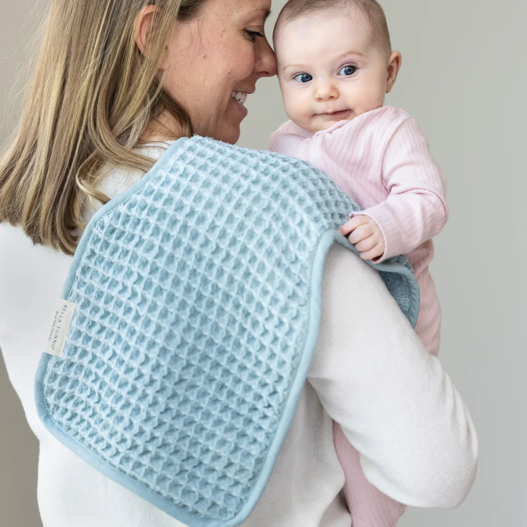 Photograph by Bella Tunno of a mother holding a baby indoors featuring a textured light blue waffle-knit burp cloth with a brand tag and a baby in a pink ribbed outfit