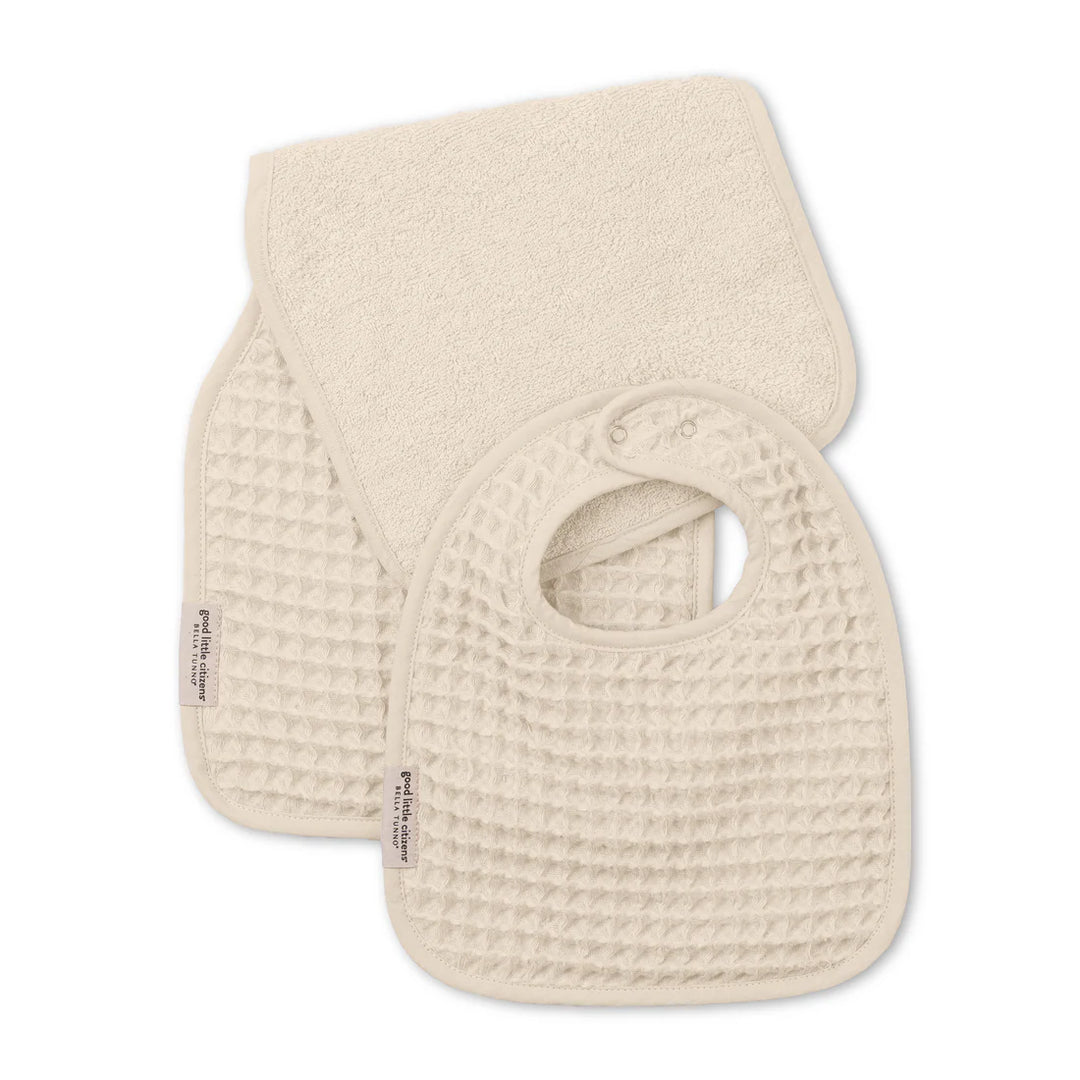 Product photograph by Bella Tunno of a cream-colored baby bib and burp cloth set featuring a distinct waffle-knit texture, snap-button closure, and small branded fabric labels against a plain white background