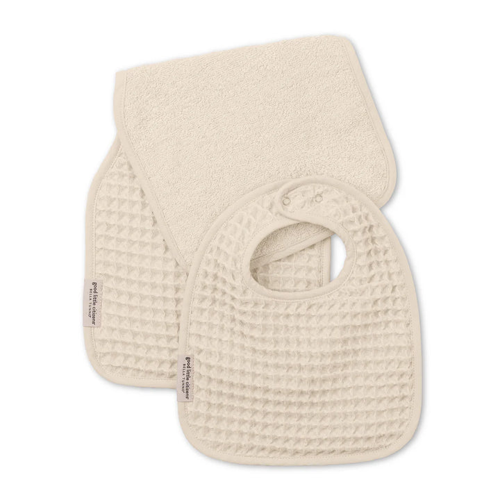 Product photograph by Bella Tunno of a cream-colored baby bib and burp cloth set featuring a distinct waffle-knit texture, snap-button closure, and small branded fabric labels against a plain white background
