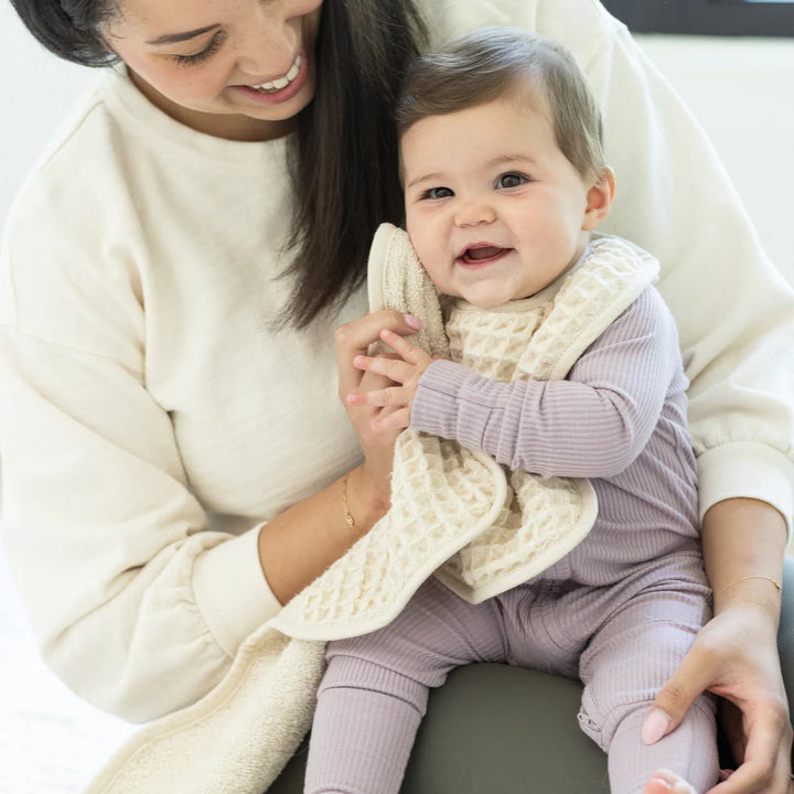 Digital photograph of a smiling mother in a white sweatshirt holding a joyful baby wearing a lavender ribbed onesie and a textured cream waffle-knit bib in a bright indoor setting