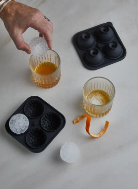 Black silicone molds with golf ball shaped ice beside two glasses of whiskey and an orange peel