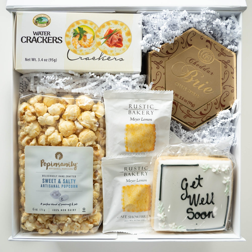 Studio photograph of a gourmet get-well gift box featuring Popinsanity artisanal popcorn, Twenty Valley Farms water crackers, a hexagonal gold box of cheddar brie, and a square hand-iced cookie with black Get Well Soon script and white floral details nestled in white crinkle paper