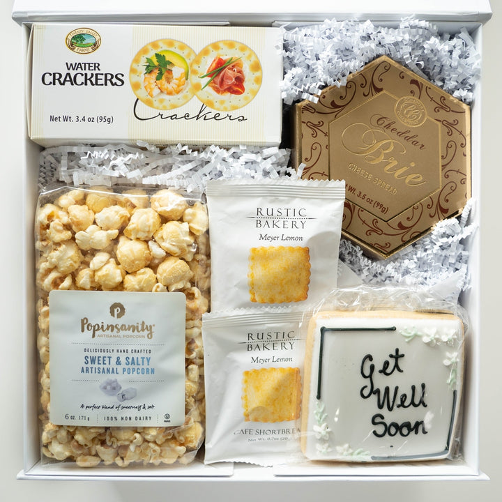Studio photograph of a gourmet get-well gift box featuring Popinsanity artisanal popcorn, Twenty Valley Farms water crackers, a hexagonal gold box of cheddar brie, and a square hand-iced cookie with black Get Well Soon script and white floral details nestled in white crinkle paper