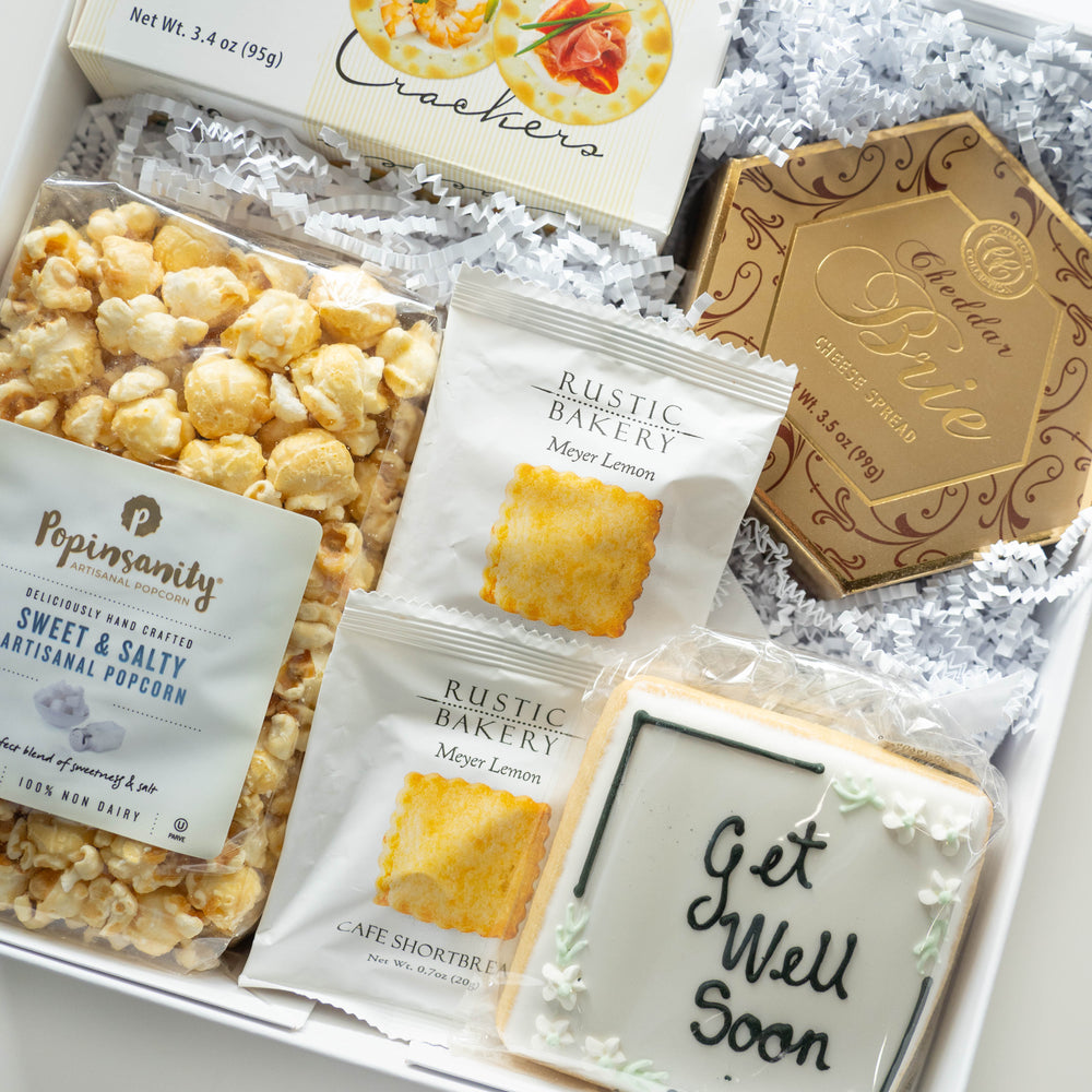 Digital photograph of a curated Get Well Soon gift box containing Popinsanity artisanal popcorn, Rustic Bakery Meyer Lemon shortbread, and a gold hexagonal box of brie spread alongside a square iced cookie with elegant black script and delicate white floral accents nestled in white shredded paper