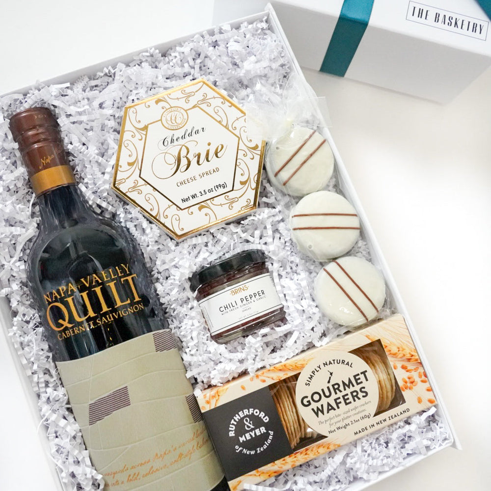 Photograph by The Basketry of a gourmet gift box featuring a Napa Valley Quilt Cabernet Sauvignon bottle, a gold-accented hexagonal brie spread, and three striped white chocolate cookies on white crinkle paper