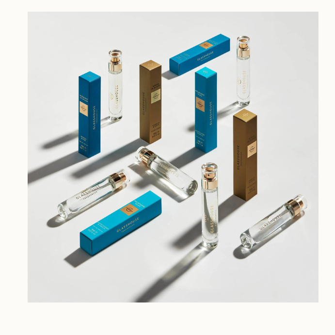Product photography by Glasshouse Fragrances featuring travel-size perfume vials and vibrant blue and bronze rectangular boxes arranged on a minimalist white surface with dramatic diagonal shadows and gold-toned spray caps