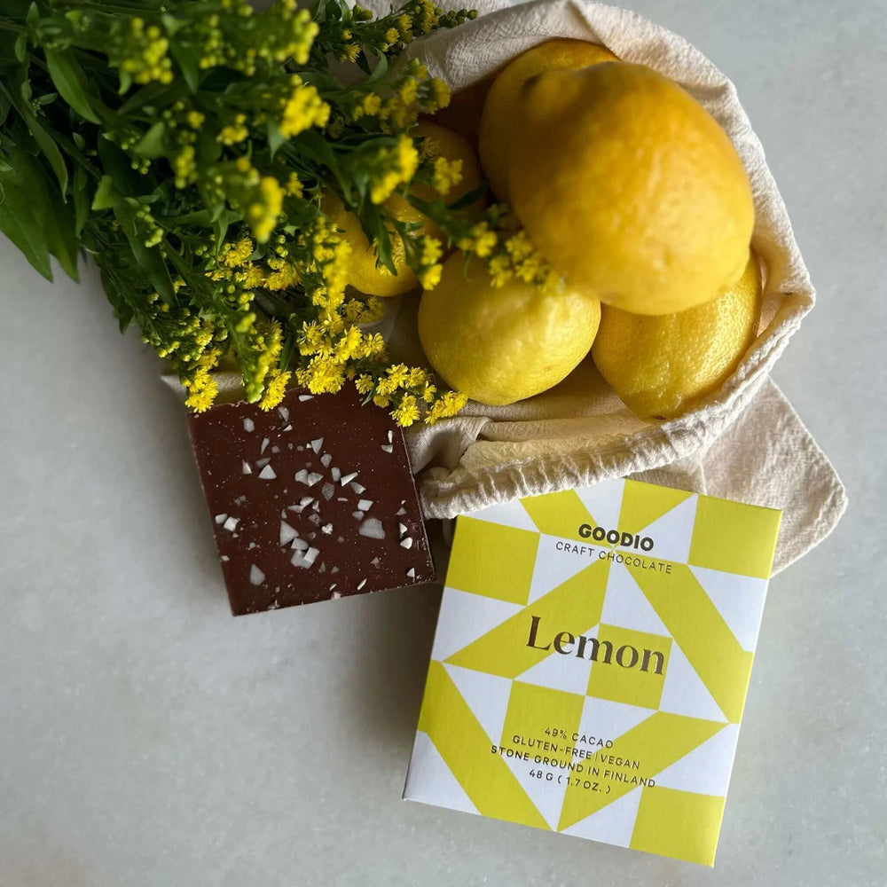 Goodio Lemon chocolate bar beside a chocolate square, lemons in a cloth bag and yellow flowers
