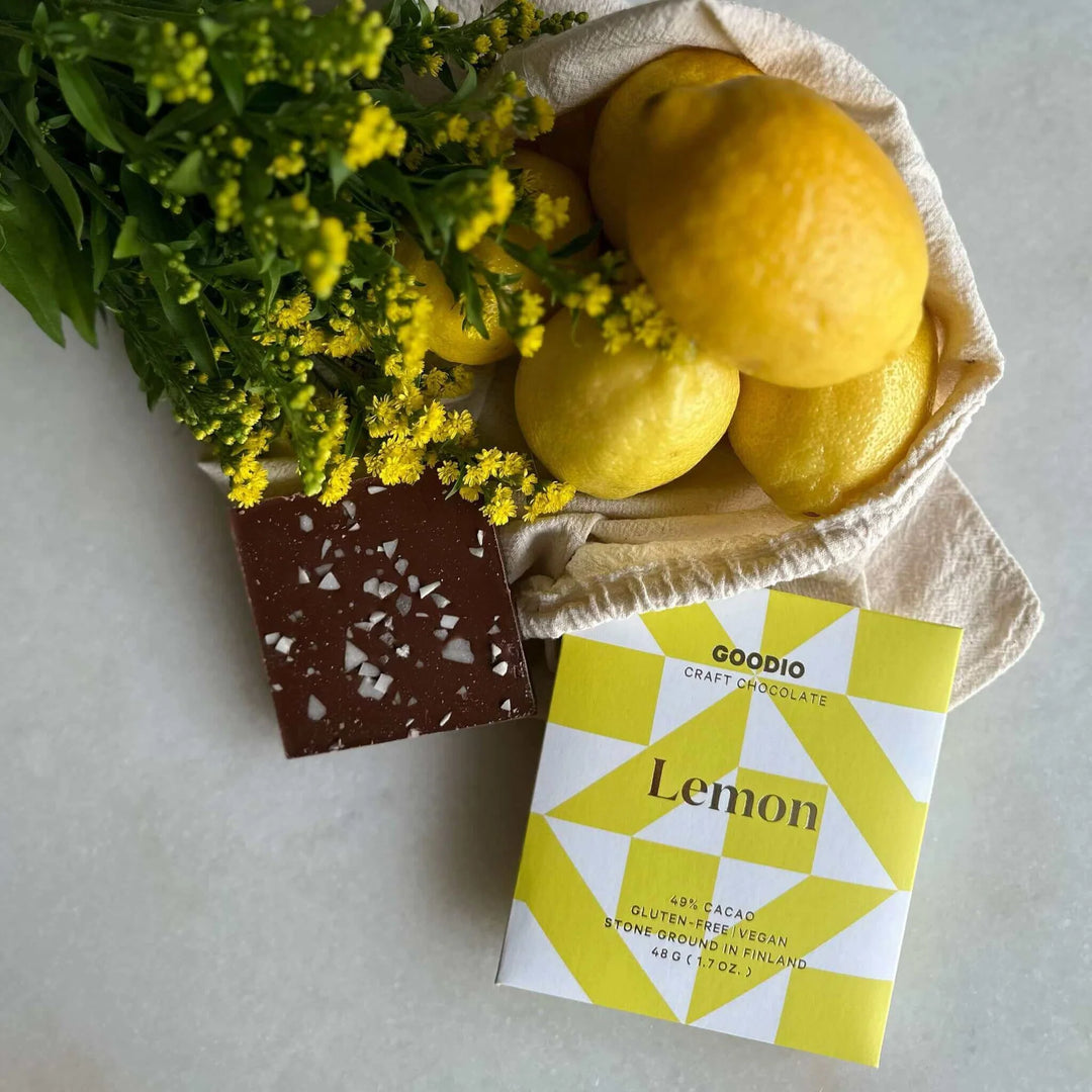 Goodio Lemon chocolate bar beside a chocolate square, lemons in a cloth bag and yellow flowers