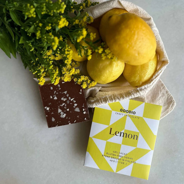 Goodio Lemon chocolate bar beside a chocolate square, lemons in a cloth bag and yellow flowers