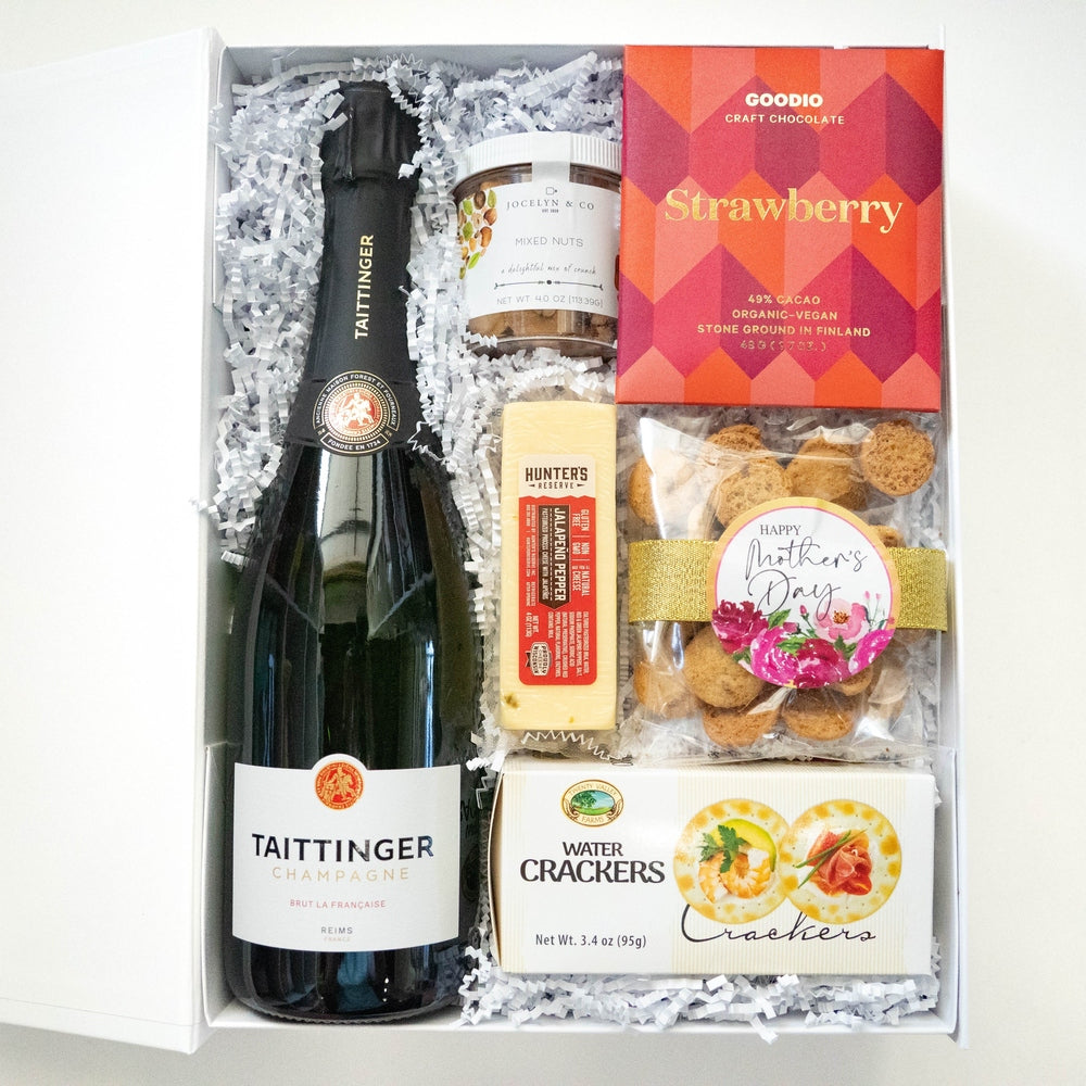 Photograph of a luxury Mother's Day gift box containing a bottle of Taittinger Champagne, a vibrant red Goodio strawberry chocolate package, and a gourmet snack bag with a pink floral sticker and gold ribbon on white shredded paper