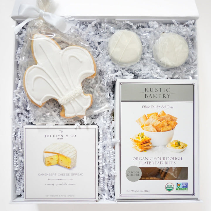 Photograph of a curated gourmet gift box by Jocelyn &amp; Co and Rustic Bakery featuring a large white fleur-de-lis iced cookie, organic sourdough flatbread bites, and Camembert cheese spread nestled in white crinkle paper with a satin ribbon bow