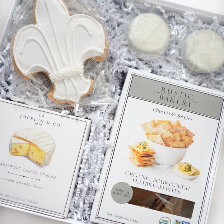 Product photography of a gourmet food gift box featuring a large white fleur-de-lis sugar cookie with decorative pearl sprinkles, Jocelyn &amp; Co Camembert cheese spread, and Rustic Bakery sourdough flatbread bites nestled in white crinkle paper shred