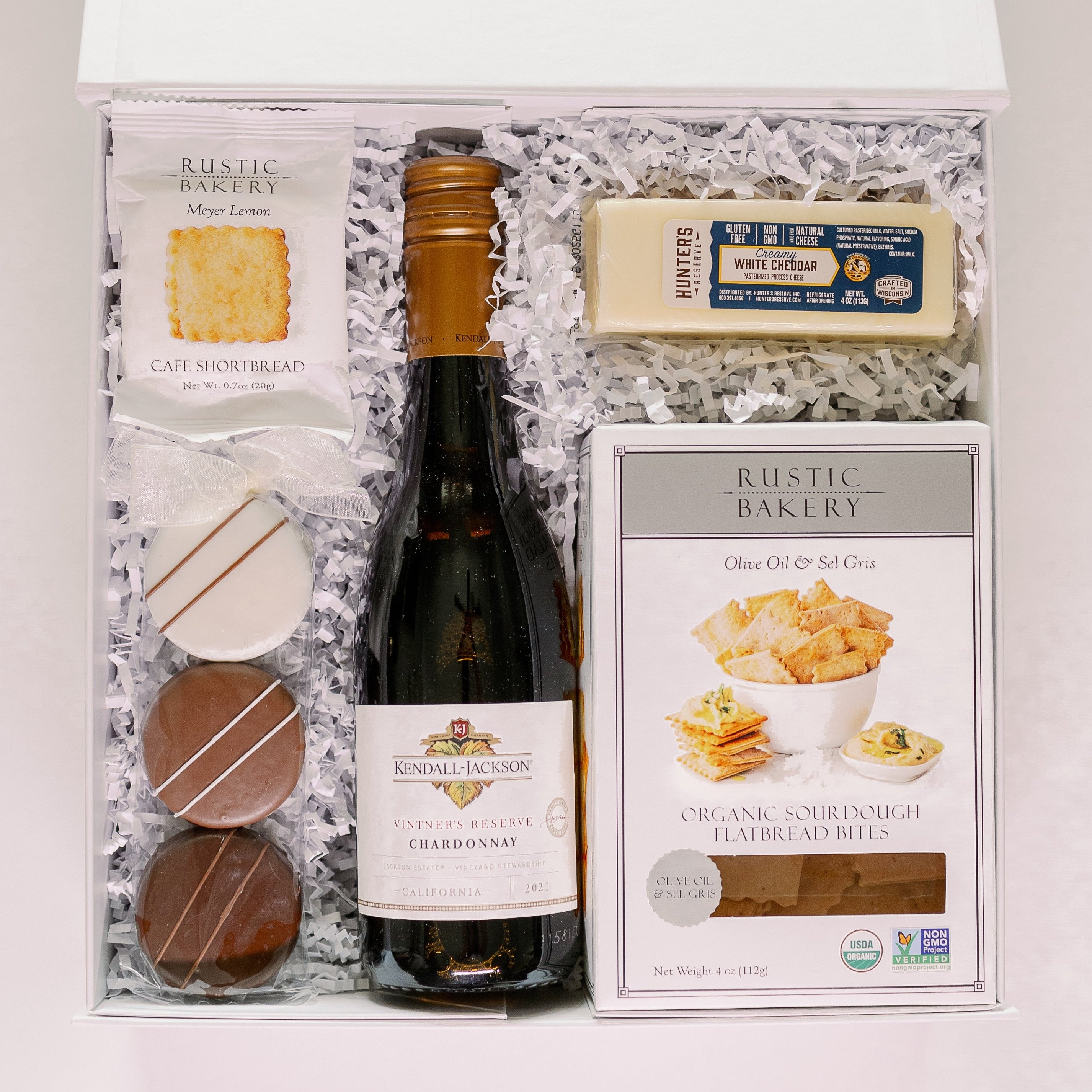 Photograph of a gourmet wine and snack gift box featuring a bottle of Kendall-Jackson Chardonnay, Hunter’s Reserve white cheddar, and Rustic Bakery snacks nestled in white crinkle-cut paper with three striped chocolate-covered cookies tied with a white bow