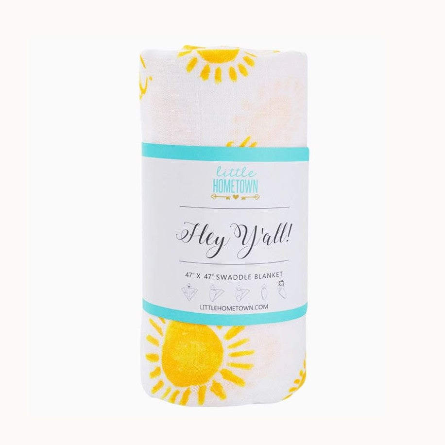 Muslin swaddle blanket by Little Hometown showcasing a pattern of hand-drawn yellow suns on white fabric wrapped in a paper sleeve with teal stripes and the handwritten phrase Hey Y'all