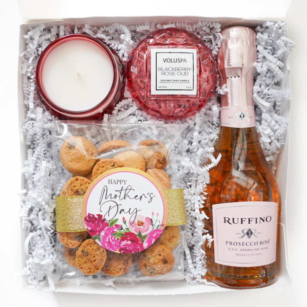 Product photograph of a Mother's Day gift box featuring a Ruffino Prosecco Rosé bottle, a red geometric Voluspa candle, and a bag of cookies decorated with a gold glitter ribbon and pink floral sticker nestled in white shredded paper
